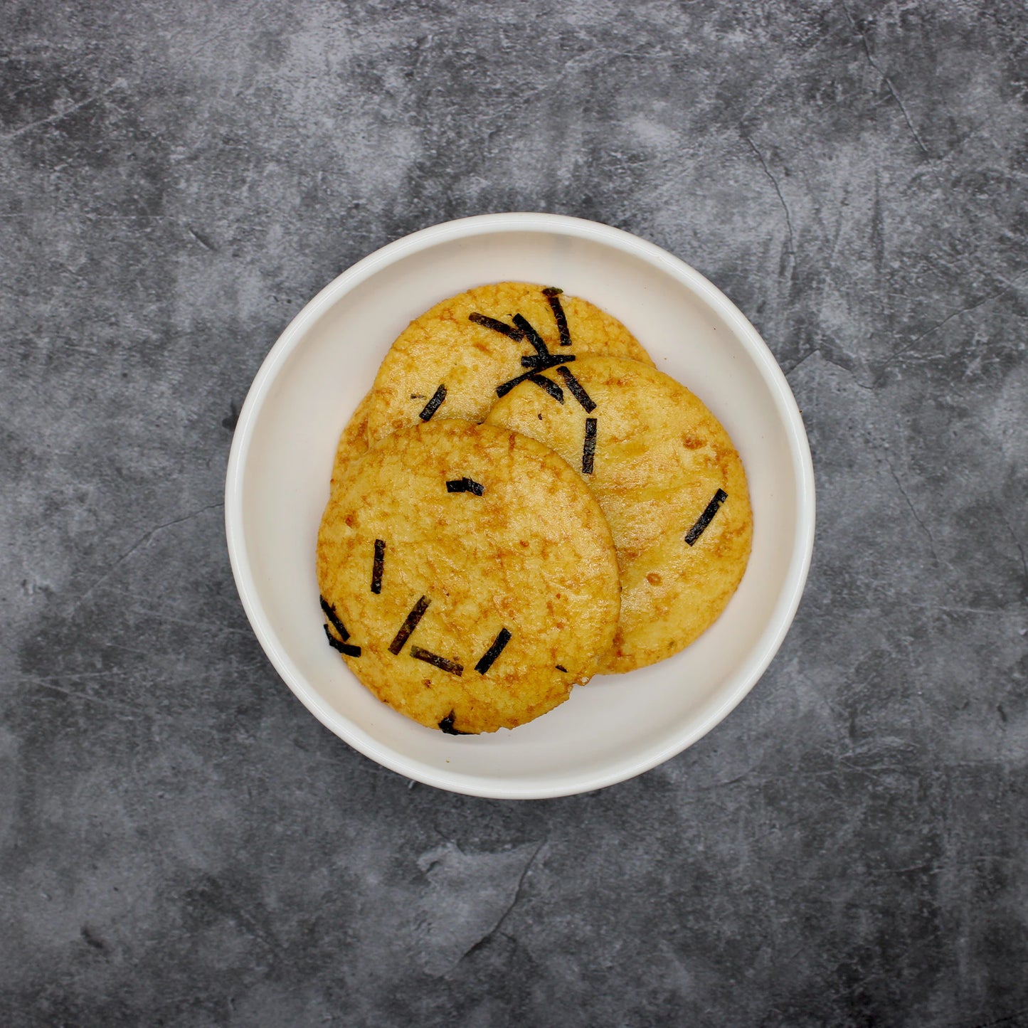 
                  
                    A white oval plate displays two Japanese Rice Crackers - Soy Sauce Senbei 110g, each topped with black seaweed strips and set on a gray textured surface for a classic savory snack.
                  
                