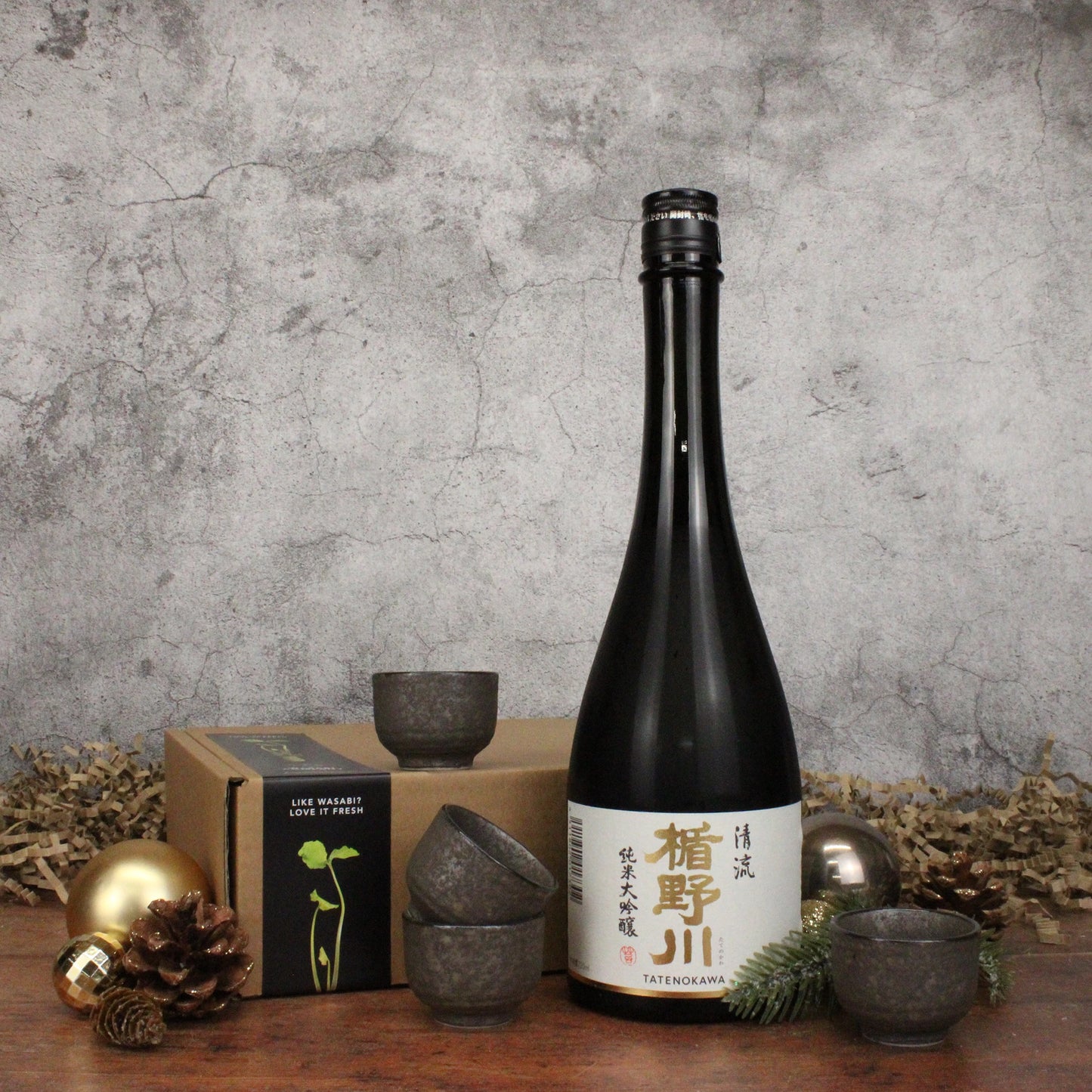 A bottle of Stream Sake with a Japanese label stands next to a set of four sake cups, a brown box, pine cone, gold ornament, and greenery on a wooden surface against a gray wall.