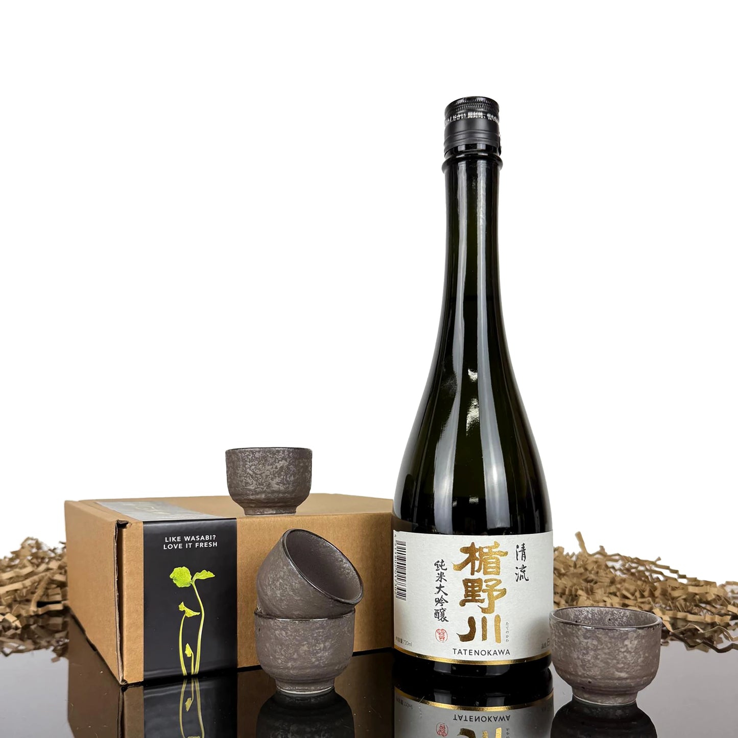 A bottle of Stream Sake with Japanese text on the label is shown next to a set of four ceramic sake cups and a brown box on a reflective surface, with shredded paper in the background.