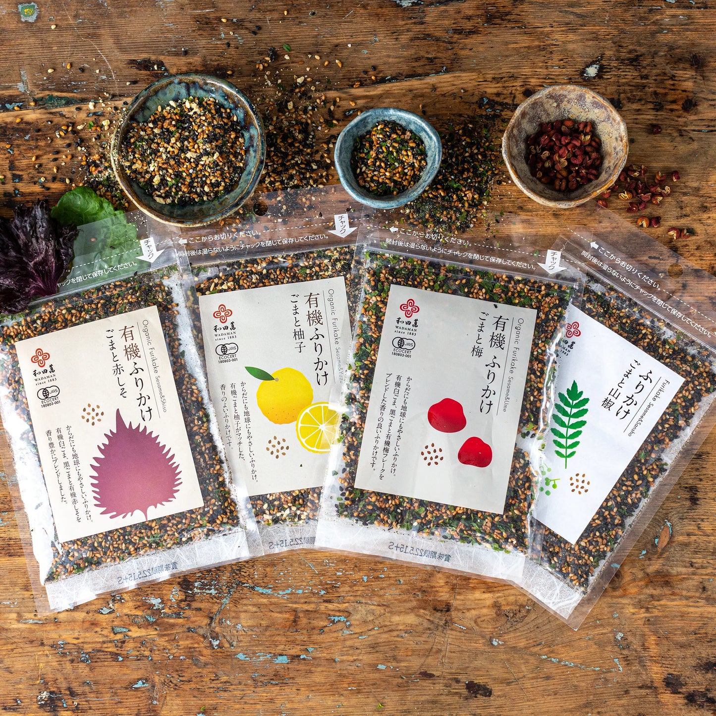 
                  
                    Four packets of Japanese furikake seasoning with different flavors are arranged on a rustic wooden table, alongside two small bowls containing seasoning.
                  
                