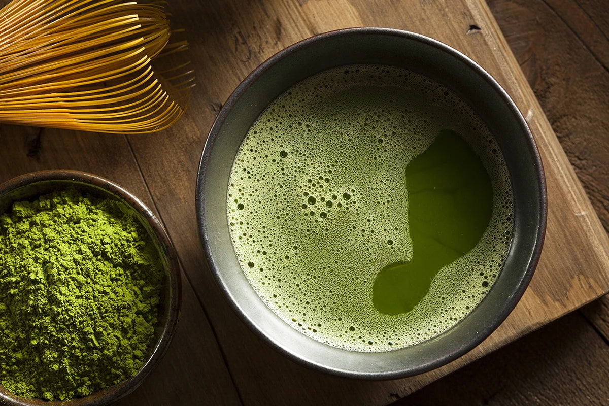 A bowl of whisked matcha tea sits on a wooden surface next to a bamboo whisk and a small bowl of matcha powder.