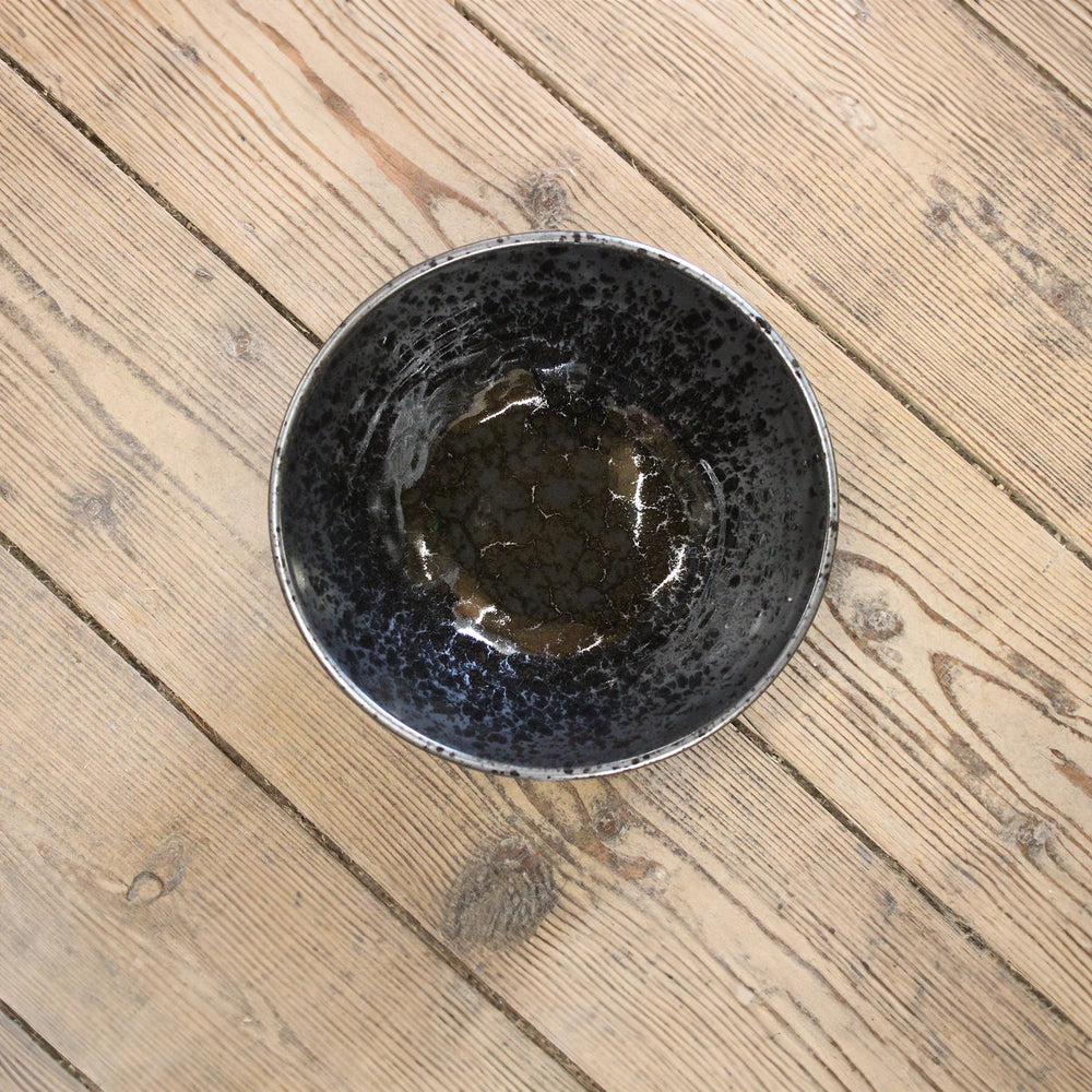 
                  
                    The Udon Bowl - Black Pearl, a Minoyaki porcelain bowl with black pearl glaze and a textured interior, sits empty on a light wood floor with visible grain—ideal as a decorative piece.
                  
                