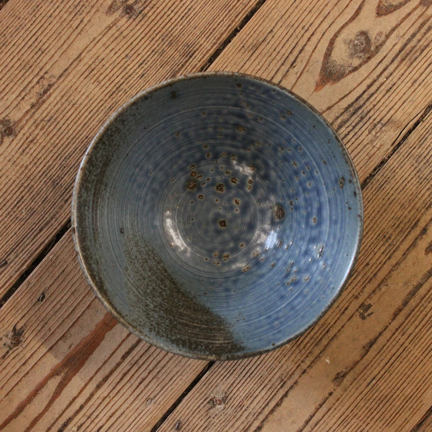 
                  
                    The Udon Bowl - Rustic Blue, a handmade Japanese bowl with a blue glazed interior and rustic finish, rests on a wooden floor showing natural grain and knots.
                  
                