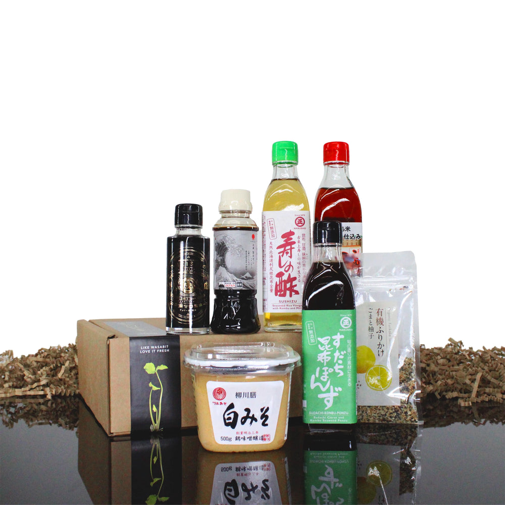 The Ultimate Vegan Japanese Cooking Collection is arranged in front of two cardboard boxes on a reflective surface, featuring ponzu sauce, white miso paste, a spice pack, and bottled vegan Japanese beverages.