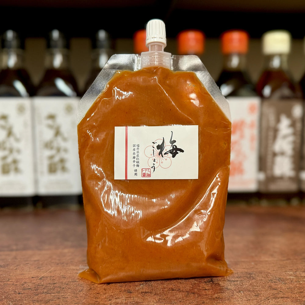
                  
                    A 300g plastic pouch of Ume Kosho – Ume Plum & Chilli Paste with a spout and Japanese label stands on a wooden surface; bottles are visible in the blurred background.
                  
                