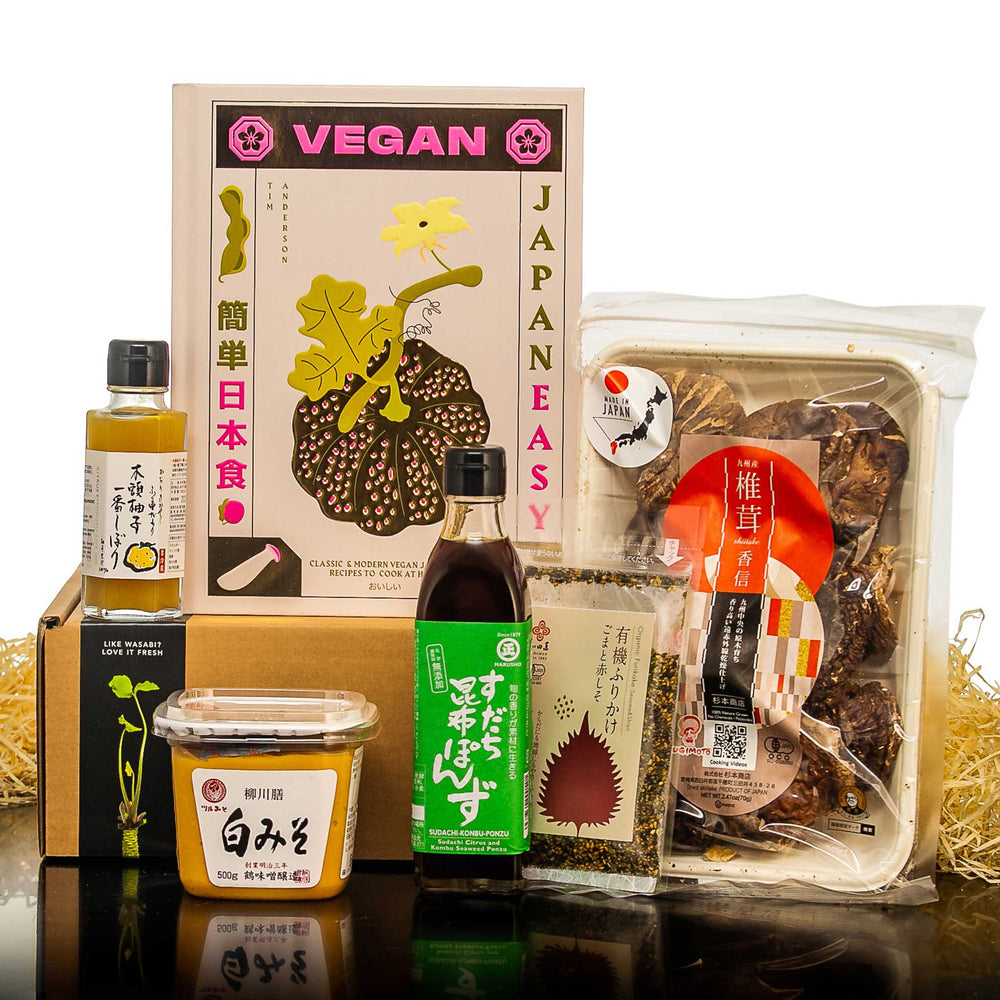 The Vegan Japanese Ingredients Set with Vegan Cook Book includes White Miso paste, Vegan Ponzu sauce, and packaged mushrooms, all displayed upright on a straw-covered surface.