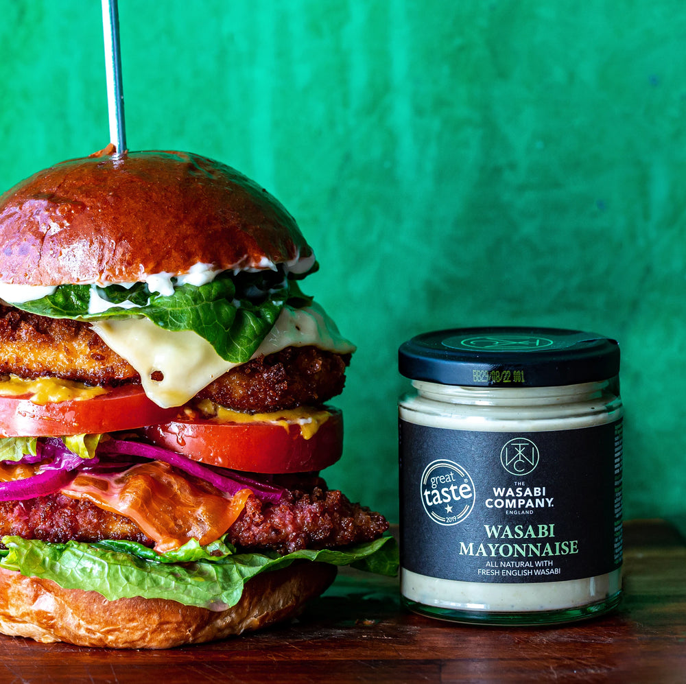 A large chicken burger with lettuce, tomato, cheese, and fried onion sits next to a jar of Wasabi Mayonnaise - 175g on a wooden surface against a green background.