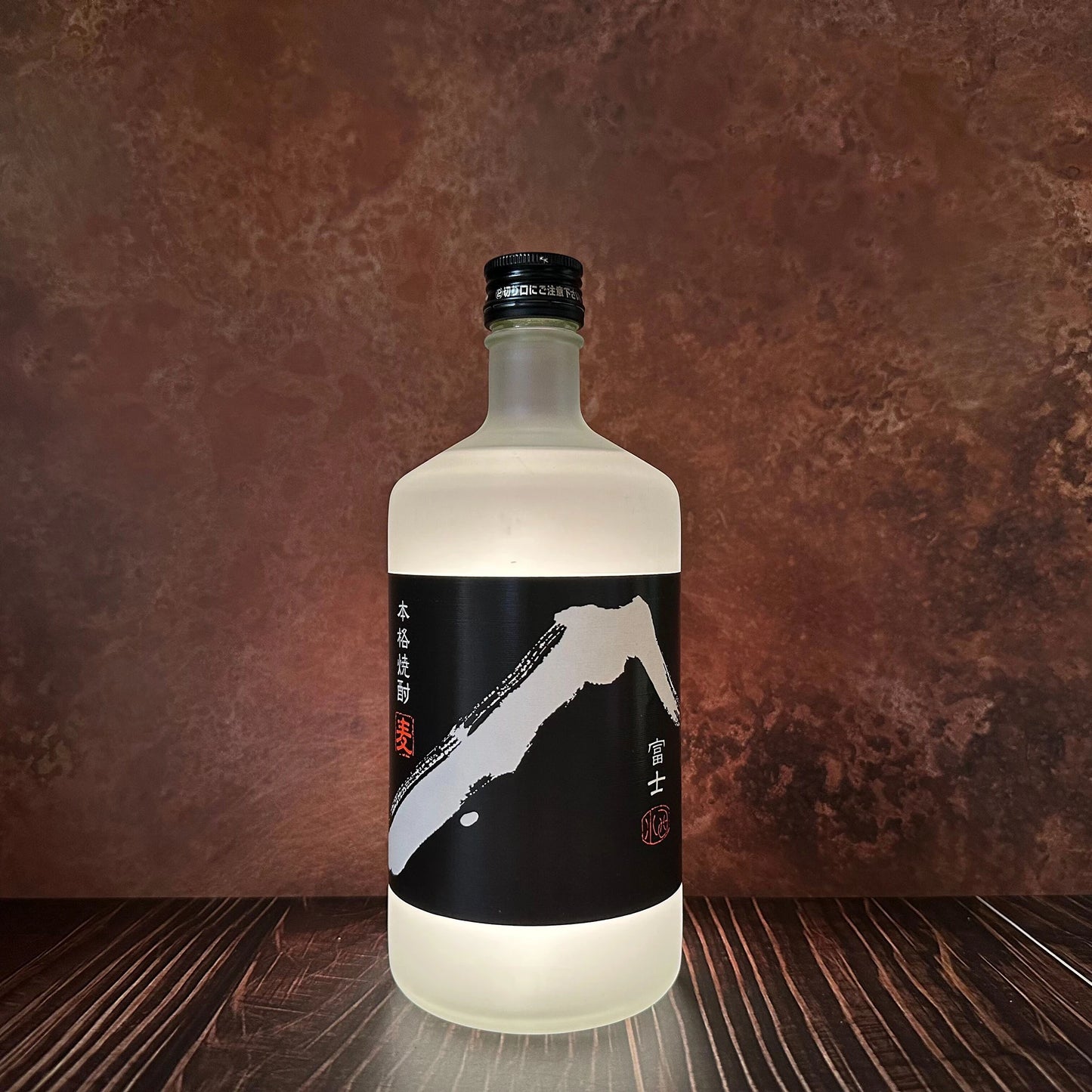 A frosted glass bottle of White Fuji Barley Shochu - 720ml with a black label featuring white abstract mountain art sits on a wooden surface, evoking purity against a brown textured background.