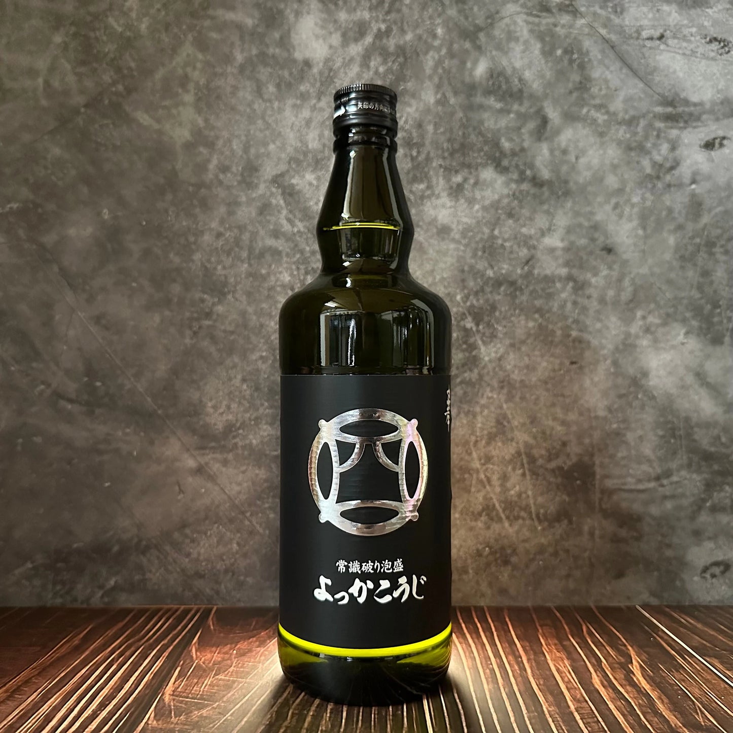 
                  
                    A dark green glass bottle of Yokka Koji Awamori from Chūkō distillery, with a black label featuring silver Japanese characters and graphics, stands on a wooden surface against a gray textured background.
                  
                