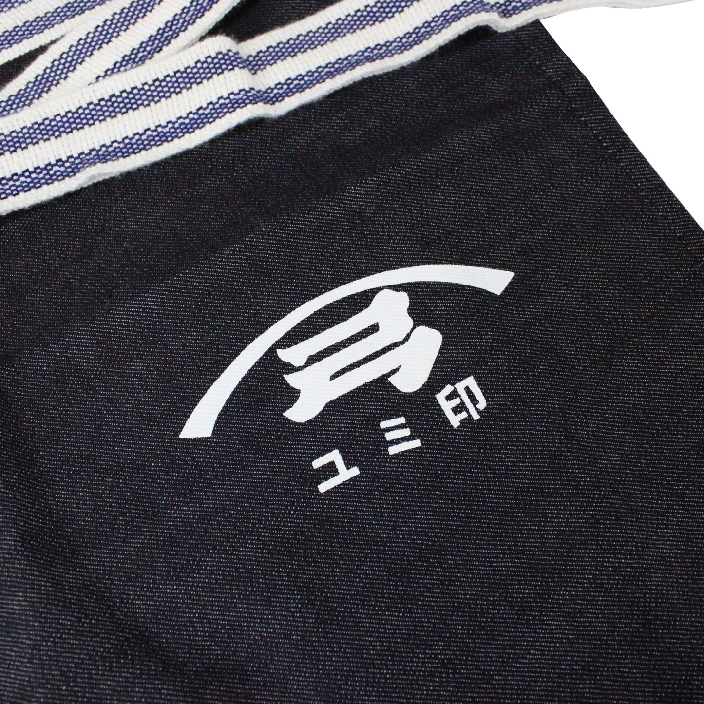 
                  
                    The Yugeta Shoyu Apron is a dark cloth apron with a white logo of Japanese characters and a curved line, partially covered by striped fabric.
                  
                