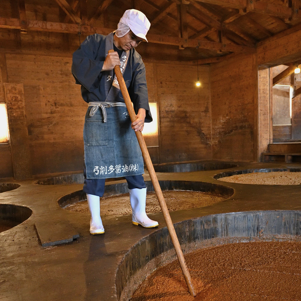 
                  
                    Wearing a Yugeta Shoyu Apron, a worker stirs a vat of fermenting ingredients in a wooden building at a soy sauce facility.
                  
                