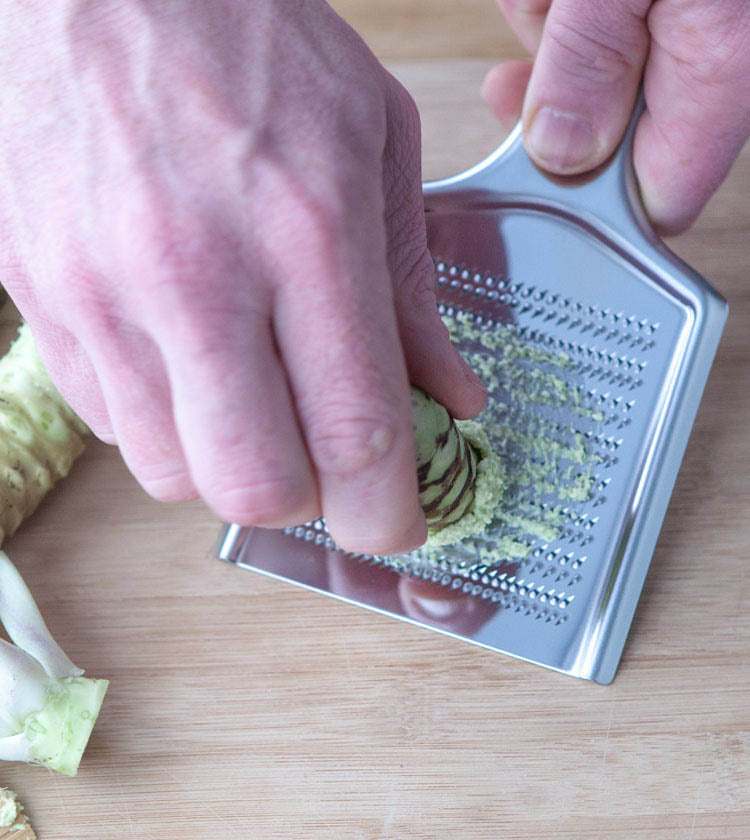 
                  
                    A person grates Individual Fresh Japanese Wasabi Rhizomes on a metal grater over a wooden surface, prized in authentic Japanese cuisine.
                  
                