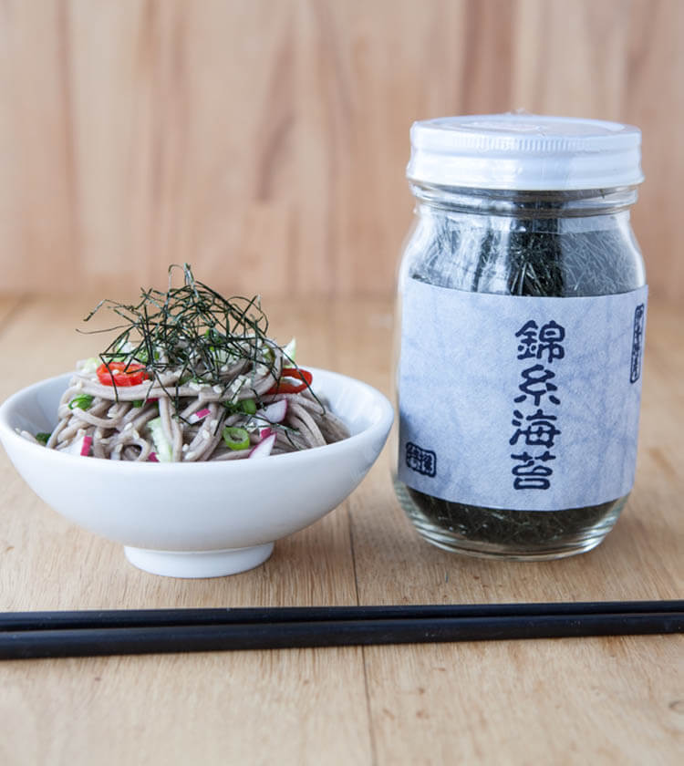 
                  
                    A bowl of soba noodles with sliced vegetables and Nori Seaweed Angels Hair Cut - 30g sits beside a jar of dried seaweed on a wooden surface, with chopsticks in front, highlighting this premium product.
                  
                