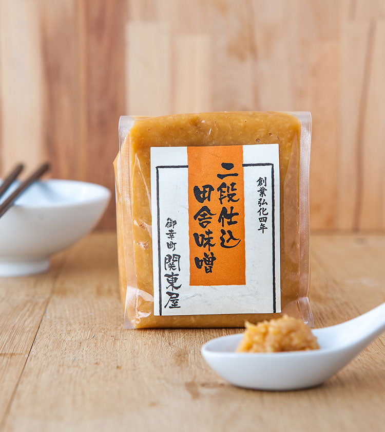 
                  
                    A sealed package of Double Fermented Rustic Miso with Japanese labels sits on a wooden surface next to a white dish holding a spoonful of the flavorful miso paste.
                  
                