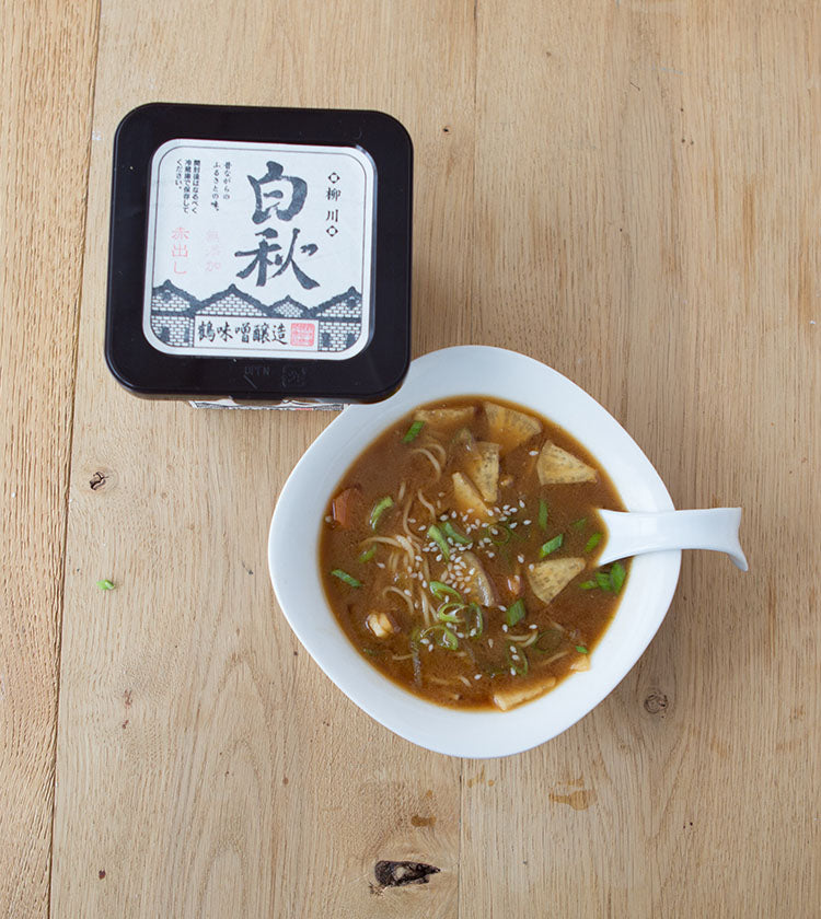 A bowl of noodle miso soup with tofu, mushrooms, and green onions sits beside a 500g Red Miso container on a wooden table, showcasing the deep umami flavor of this essential ingredient.