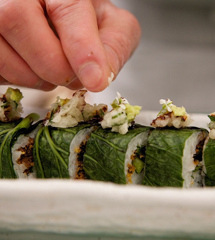 
                  
                    A hand adds small toppings to sushi rolls wrapped in English Wasabi Baby Leaf - 30g, presented on a white rectangular plate with a fresh salad garnish and vibrant English-grown wasabi.
                  
                