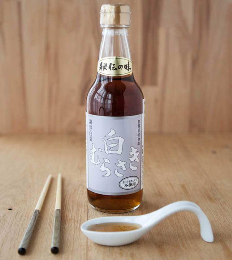 
                  
                    A bottle of Shiro Murasaki, White Soy Sauce with a white label sits on a wooden surface beside chopsticks and a white ceramic spoon filled with soy sauce.
                  
                