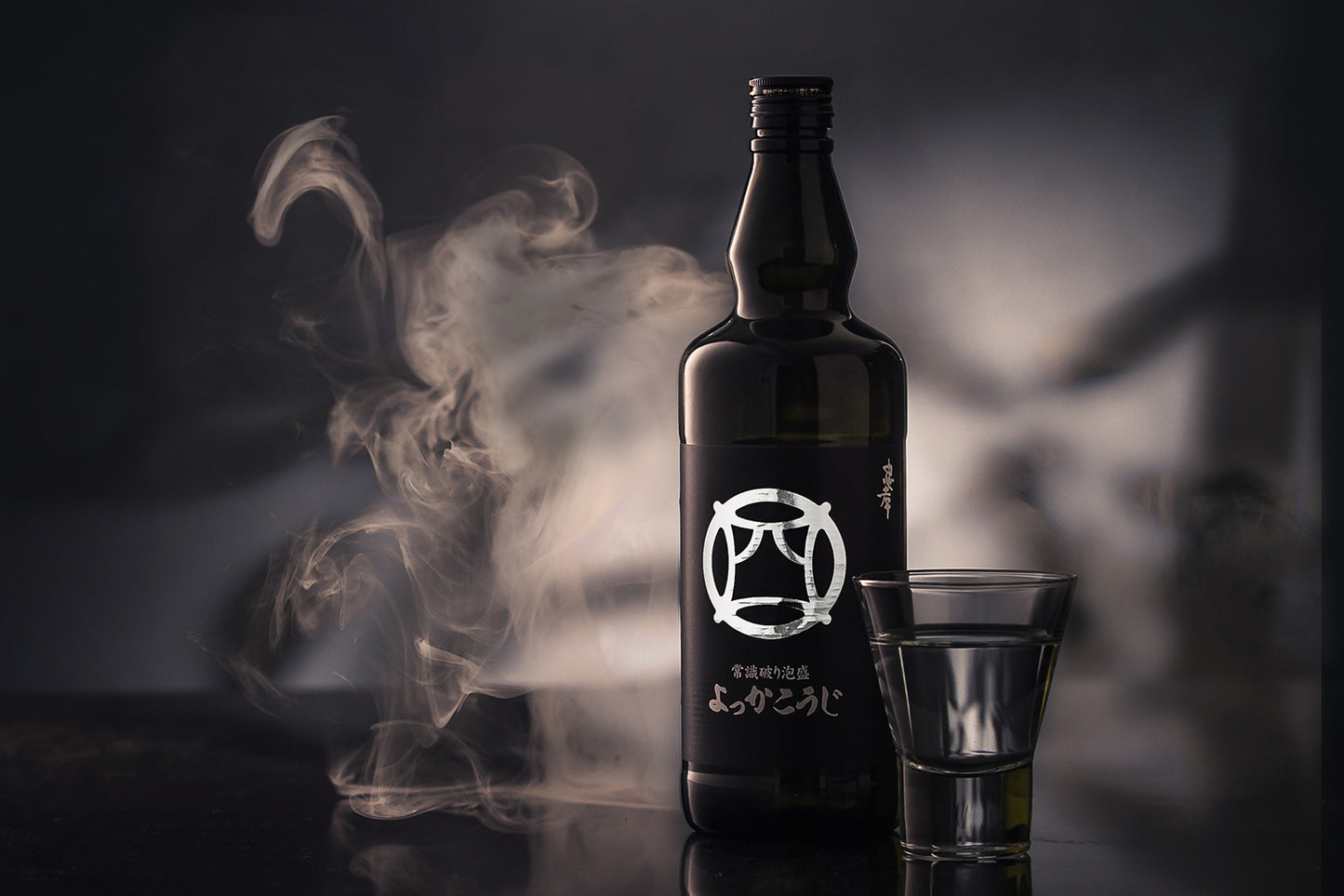 
                  
                    A dark glass bottle of Yokka Koji Awamori from Chūkō Distillery, labeled in Japanese, stands next to a clear glass of Awamori on a reflective surface with drifting smoke, capturing the spirit of Okinawa.
                  
                
