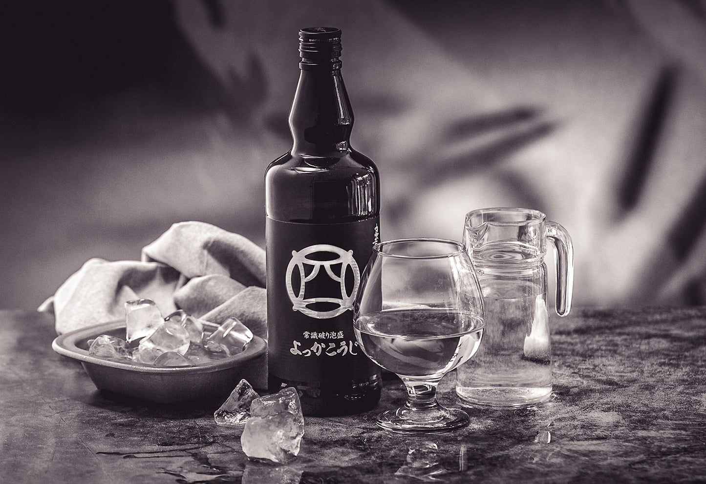 
                  
                    A bottle of Yokka Koji Awamori from the Chūkō distillery, a glass of clear Awamori, a jug of water, and a bowl of ice cubes rest on a marble surface.
                  
                