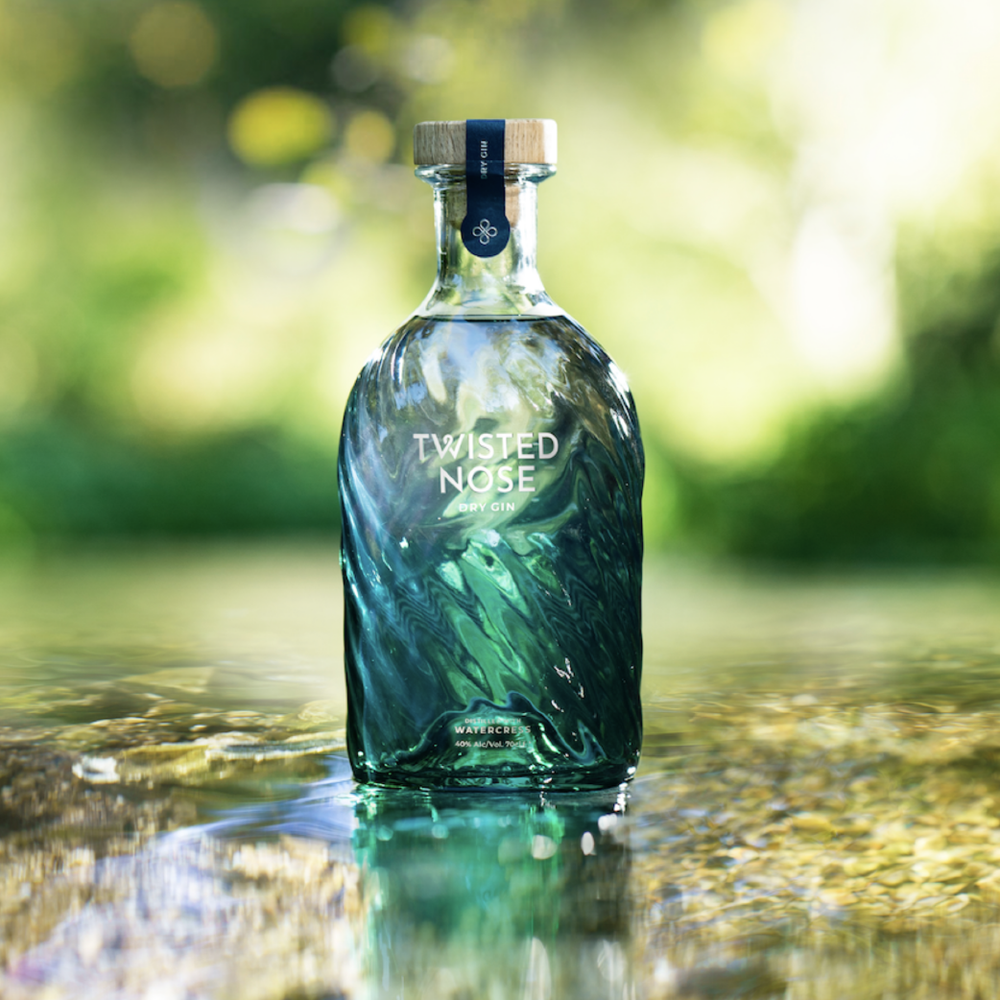 A clear glass bottle of Watercress Gin - 70cl stands upright in shallow water, set against a softly blurred green natural background.