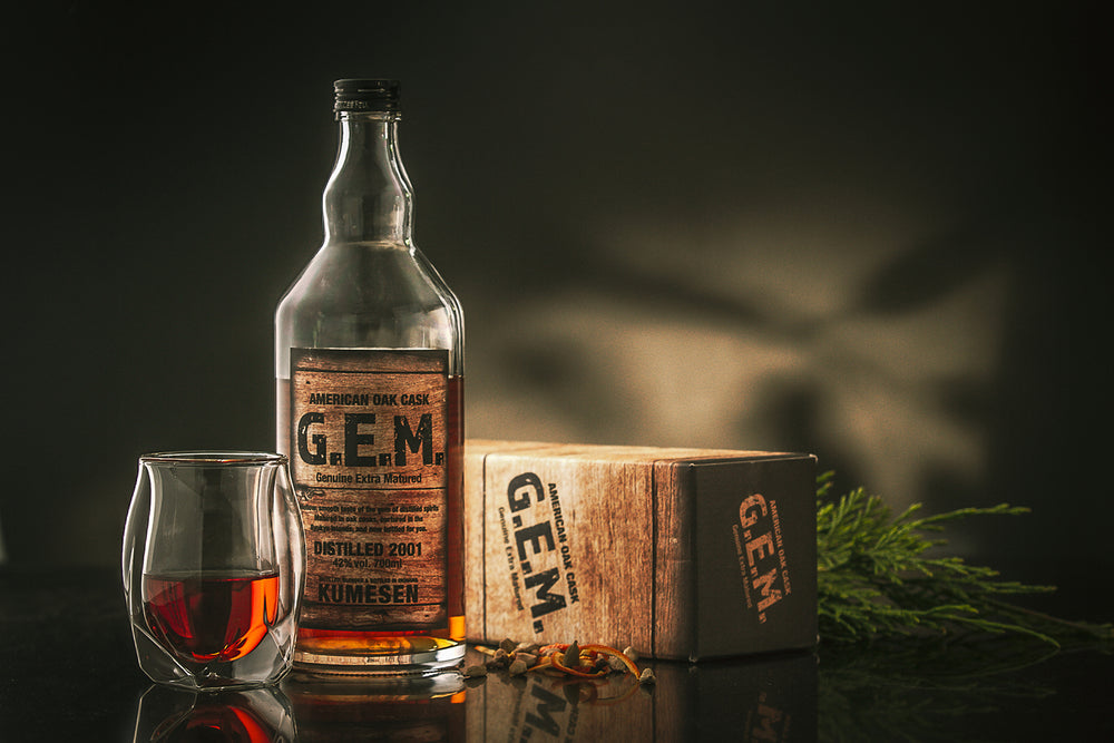 
                  
                    A bottle of Kumesen G.E.M. Awamori 10 Year Aged - 700ml, a glass of the spirit, a labeled box, and dried spices are arranged on a dark surface with greenery in the background.
                  
                