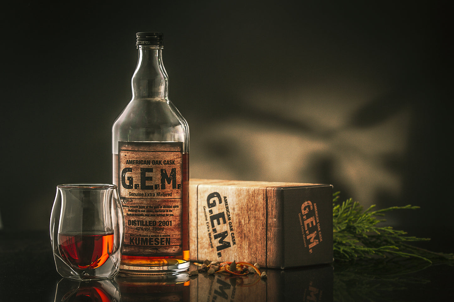 
                  
                    A bottle of Kumesen G.E.M. Awamori 10 Year Aged - 700ml, a glass of the spirit, a labeled box, and dried spices are arranged on a dark surface with greenery in the background.
                  
                