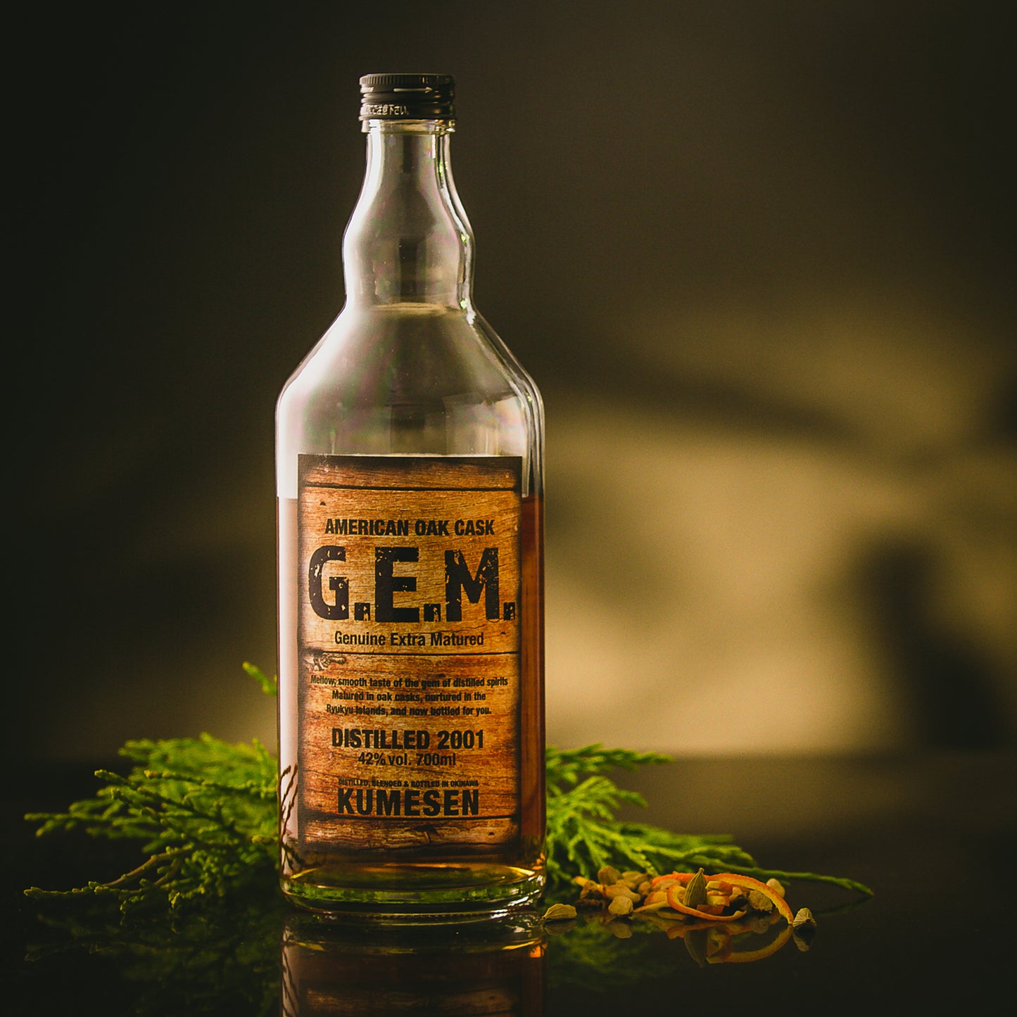 
                  
                    A bottle of Kumesen G.E.M. Awamori 10 Year Aged - 700ml rests on a dark surface with greenery and dried herbs, reflecting its oak-matured depth and refined character.
                  
                