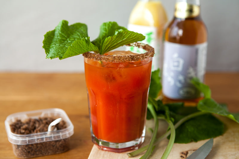 
                  
                    A glass of red cocktail with ice and a spiced rim, garnished with leafy greens, sits on wood—its unique flavor enhanced by a splash of Shiro Murasaki White Soy Sauce, with bottles and seasoning nearby.
                  
                