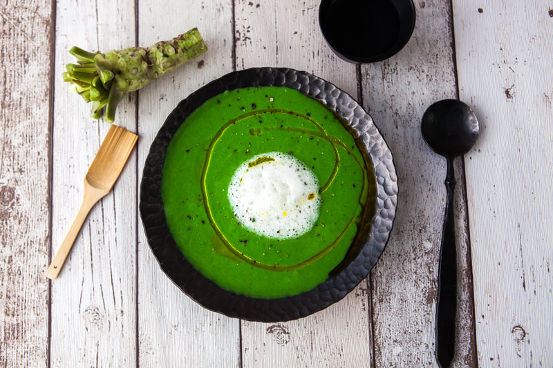 
                  
                    A black bowl of green soup with white foam and oil sits on a rustic table next to a spoon, wooden spatula, cup, and Small Fresh English Wasabi Rhizomes 200g that enhance the dish’s natural sweetness.
                  
                