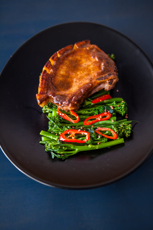 
                  
                    A cooked pork chop is served over broccolini and sliced red chili peppers, finished with a sprinkle of Sesame Seeds with Wasabi, all presented on a black plate.
                  
                