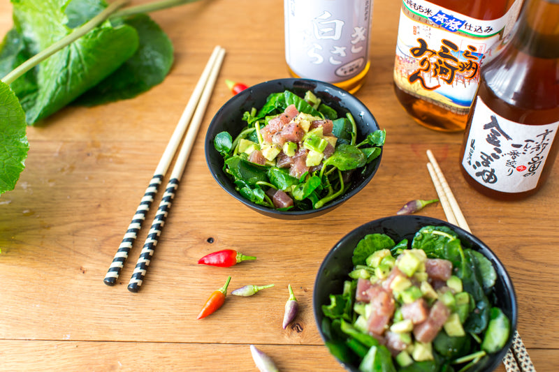 
                  
                    Two salad bowls with diced fish and avocado are served among chopsticks, assorted peppers, leafy greens, and Japanese drinks—featuring Shiro Murasaki White Soy Sauce—on a wooden table.
                  
                