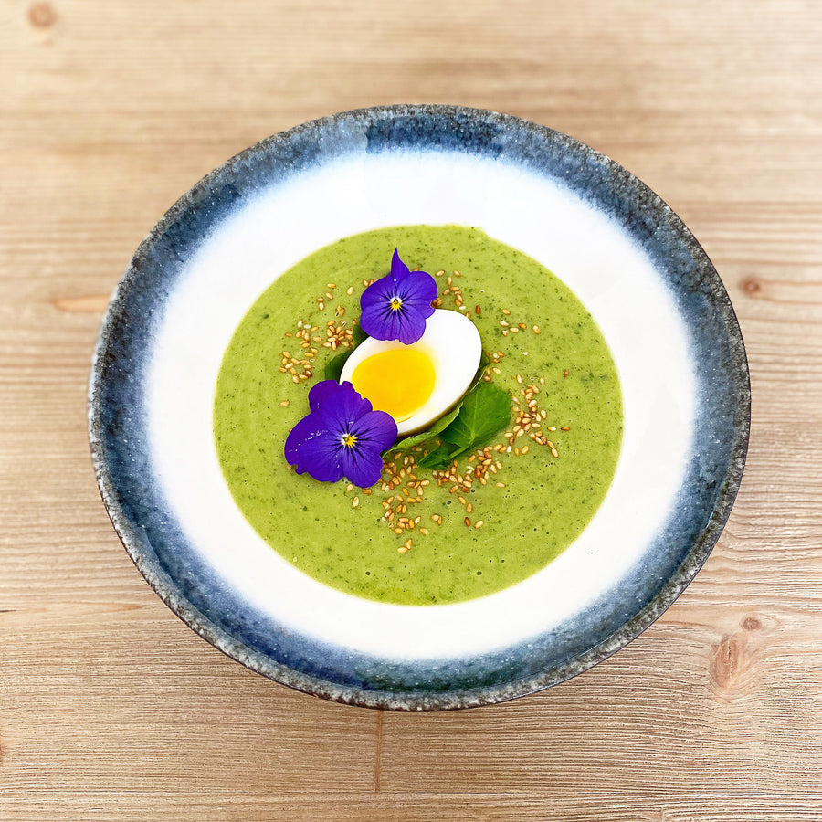 
                  
                    A vibrant green soup made with Fresh Watercress, garnished with half a boiled egg, edible purple flowers, and sesame seeds, presented on a wooden surface.
                  
                