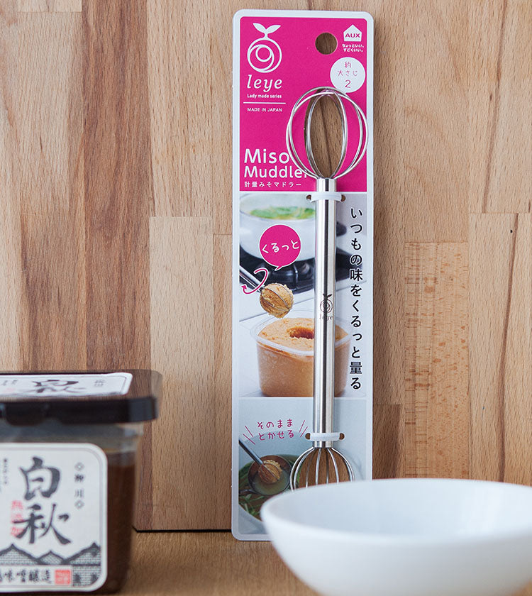 The Miso Muddler stands upright against a wooden background, with a bowl, a tub of miso paste, and a whisk placed nearby.