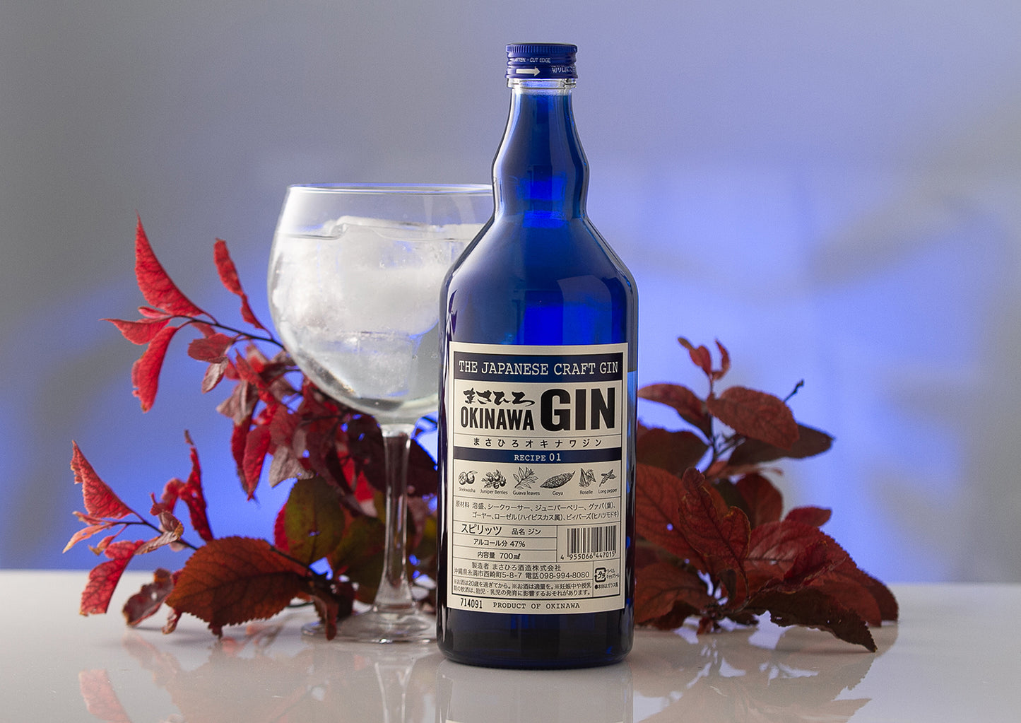 
                  
                    A bottle of Okinawa Gin - 700ml, crafted with Okinawan botanicals and Awamori, is placed next to a glass of gin on ice, set against vibrant red leaves and a softly blurred blue backdrop.
                  
                