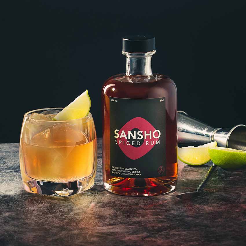 
                  
                    A glass of cocktail with a lime wedge is beside a bottle of Sansho Spiced Rum - 50cl, infused with Japanese Sansho Berries. Lime slices and a metal jigger are on the dark surface in the background.
                  
                