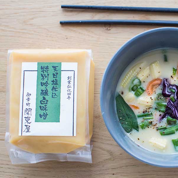 
                  
                    A block of Shiro White Miso beside a bowl of vegetable soup and black chopsticks on a light wooden surface, highlighting the importance of fermentation in Japanese cuisine.
                  
                