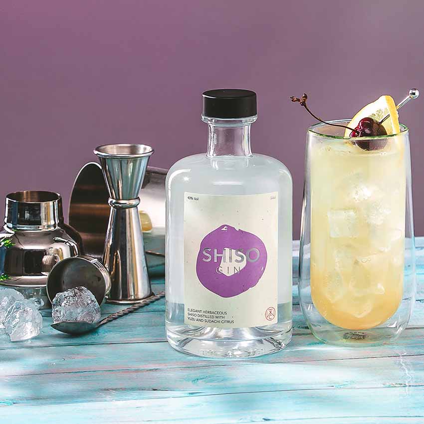 
                  
                    A 50cl bottle of Shiso Gin with purple shiso, a cocktail in a tall glass with ice, lemon and cherry garnish, metal bar tools, and ice cubes on a blue wooden surface against a purple background.
                  
                