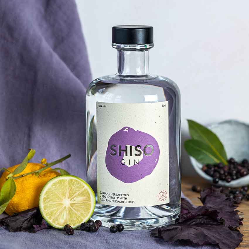 A bottle of Shiso Gin - 50cl sits on a surface surrounded by a halved lime, yuzu, purple shiso leaves, green leaves, and juniper berries.