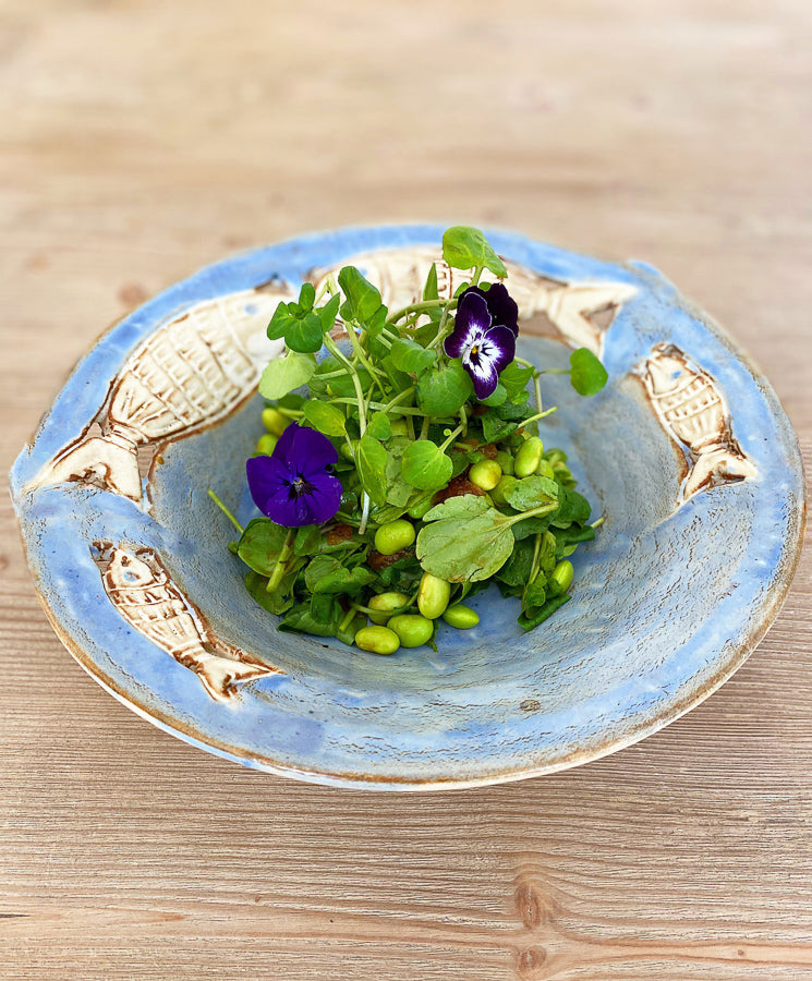 
                  
                    Fresh Watercress is served in a ceramic bowl with fish designs, accompanied by leafy greens, edamame, and two purple edible flowers, all arranged on a wooden surface.
                  
                