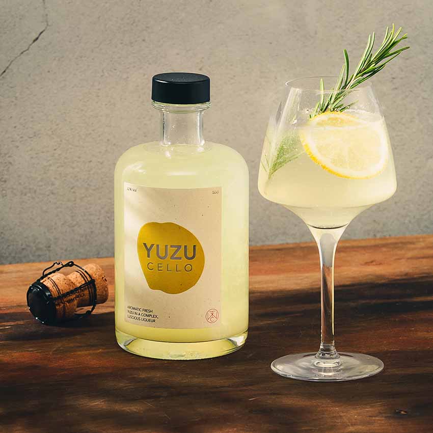 
                  
                    A bottle of Yuzucello - 50cl stands beside a glass of yellow cocktail garnished with a lemon slice and rosemary, highlighting its citrus flavors and aromatic blend on a wooden surface.
                  
                