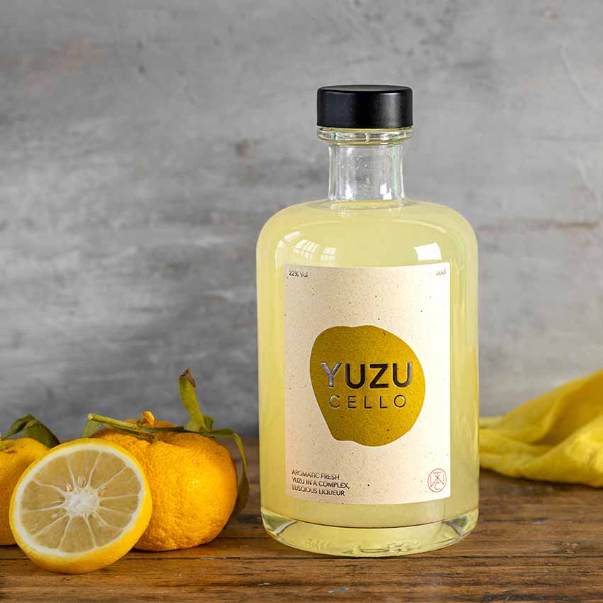 A bottle of Yuzucello - 50cl stands on a wooden surface beside a whole yuzu fruit and sliced lemon, emphasizing its aromatic citrus blend against a gray background.